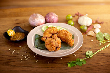 A top-down view of crispy Asian-style fried chicken drumsticks on a wooden table, accompanied by various spices such as cilantro, chili sauce, green onions, garlic, lime, and dried chili peppers.