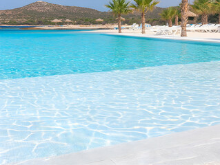 Fototapeta premium Crystal-clear infinity pool water reflects sunlight near a beach with palm trees and a mountain background, offering a serene summer escape.