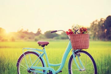 Fototapeta premium A vintage-style turquoise bicycle with a wicker basket filled with colorful flowers rests in a lush green field during a warm sunset, evoking a peaceful and serene atmosphere.