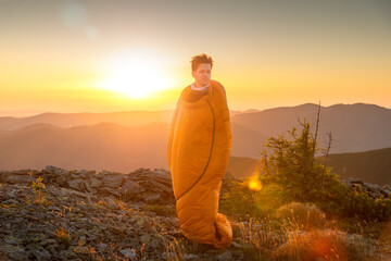Obraz premium A young man with tousled hair stands wrapped in an orange sleeping bag on a rocky ridge at sunrise, gazing into the distance as golden light illuminates the mountains.