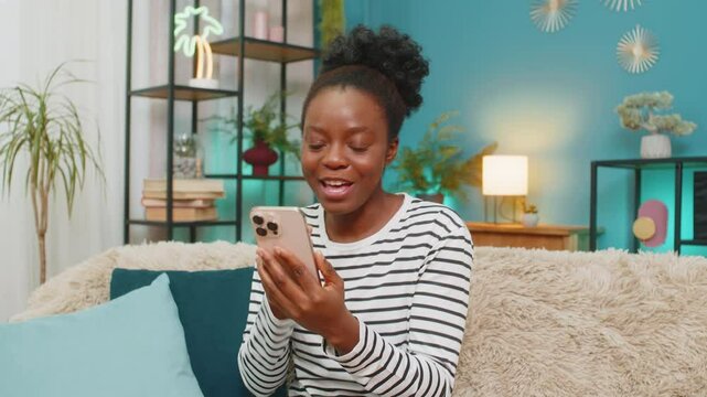 African American woman celebrating good news on smartphone. Sitting at home sofa, joyful expression, happy emotions, receiving message or winning in lottery, engaging indoor atmosphere, excited mood