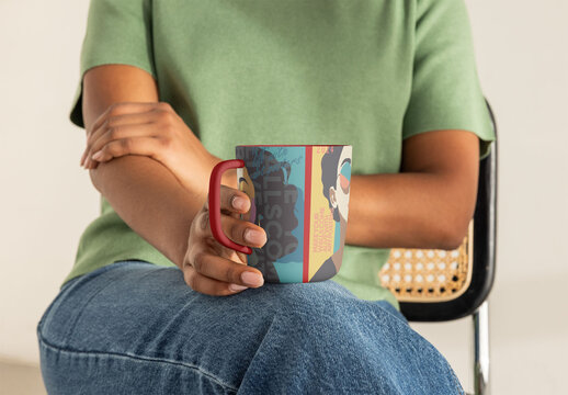 Handmade ceramic mug with handle held in female hand while seated on a chair. PSD template mockup
