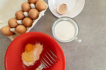 A red mixing bowl contains flour and a cracked egg, with a whisk resting inside.