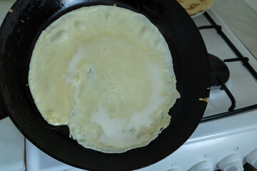 A thin crepe is cooking in a black frying pan on a gas stove.