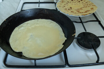 A thin crepe is cooking in a black frying pan on a gas stove, with a stack of prepared crepes on a plate in the background. 