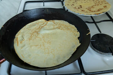 A thin crepe is cooking in a black frying pan on a gas stove, with a stack of prepared crepes on a plate in the background. 