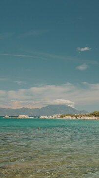 Vertical Screen: Tourists enjoying crystal clear turquoise waters, sunbathing and swimming along picturesque Greek coastal landscape under bright summer sky