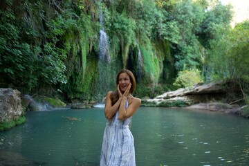 Fototapeta premium A joyful woman in a white dress stands by a scenic river with a waterfall in the background, expressing excitement and happiness in a lush natural setting.