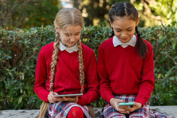 Two schoolgirls using electronic devices sitting on a bench in a park