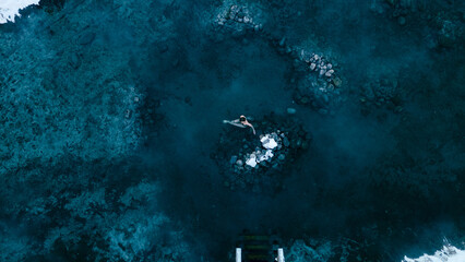 Aerial view of a lone woman relaxing in a stunning thermal spring surrounded by snowy landscapes in...