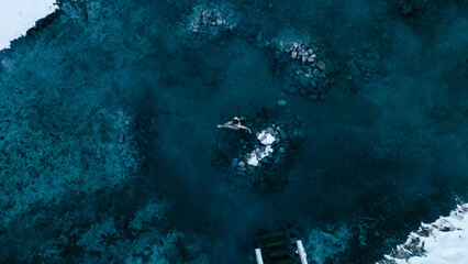 Aerial view of a lone woman relaxing in a stunning thermal spring surrounded by snowy landscapes in...