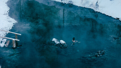 Aerial view of a lone woman relaxing in a stunning thermal spring surrounded by snowy landscapes in...