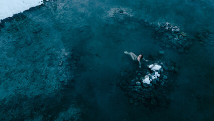 Aerial view of a lone woman relaxing in a stunning thermal spring surrounded by snowy landscapes in...