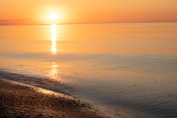 Naklejka premium Dawn on the seashore. Calm golden water and sparkling golden stones on the shore, the sun rising over the horizon, a path of sunlight on the water, everything in a golden warm light