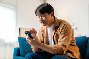 Cheerful Asian male engaging with smartphone, scrolling content, streaming video, gaming, networking, ordering food while relaxing at home