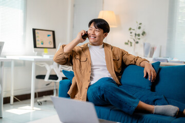 Happy young asian man talking on mobile phone while relaxing on sofa in home office
