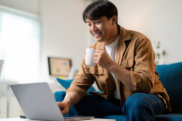Smiling asian freelancer working remotely, sipping coffee beside laptop at home workspace