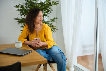 Relaxed Woman in Yellow Shirt Sitting by Window with Smartphone