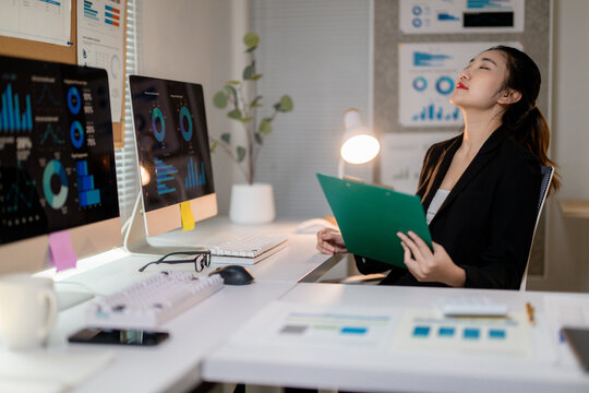 Asian businesswoman relaxing while working with financial data on computer and clipboard in office at night
