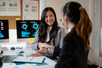 Two happy businesswomen shaking hands after successful meeting in office with charts and graphs on computer screens