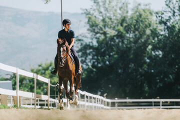 A man rides a horse on a fenced equestrian track surrounded by lush greenery. It's a sunny day,...