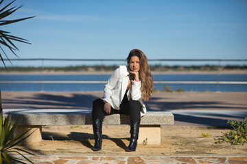 Young, beautiful woman, brown hair, wearing a white jacket, looking at the camera, with her hand on her chin, sitting facing forward on a bench, lonely and sad, receiving the sun's rays at dawn.