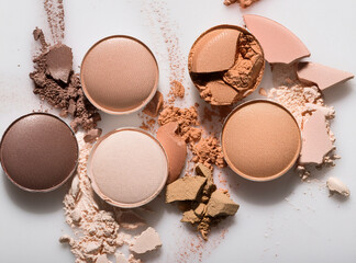 Generated image shows a collection of makeup powders in various shades of crushed eyeshadow palettes in various shades, including light, medium, and dark brown tones, scattered on a white surface