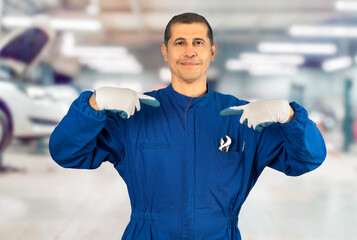 Middle age handsome man wearing mechanic blue uniform looking confident with smile on face, pointing oneself with fingers proud and happy at repair shop.