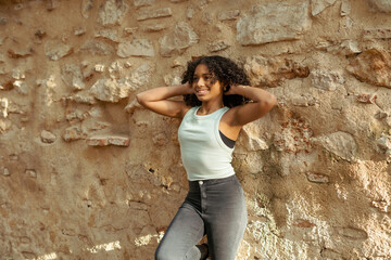 Young woman posing with hands on her curly hair against an old wall
