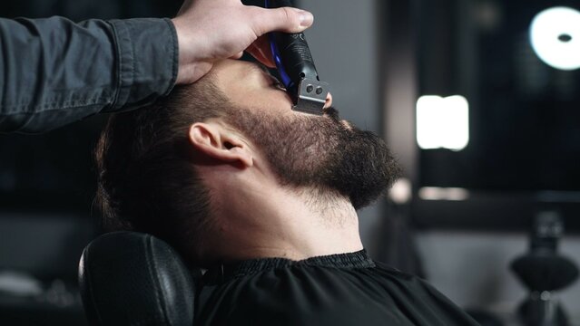 Professional barber trimming client's beard with electric razor, delivering precise styling within contemporary grooming salon setting