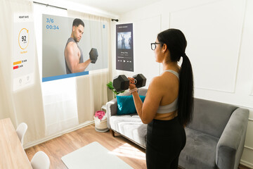 Woman wearing AR glasses, tracking fitness routine on virtual display while strength training with dumbbells in home workout space