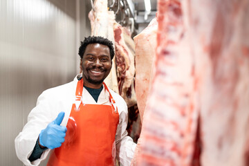 Professional butcher holding thumbs up and working with raw meat in slaughterhouse.