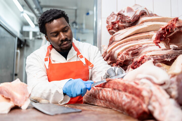 Professional African American butcher slicing pork meat inside slaughterhouse.