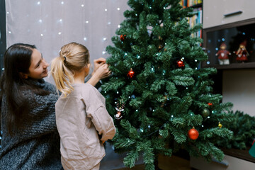 Little girl stands watching her mother hanging balls on the Christmas tree. Back view