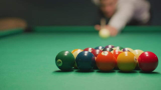 Blurred figure in light shirt and dark trousers leans over green pool table, gripping cue stick with deep concentration. Eyes locked on balls, posture steady, creating tension in dimly lit game room