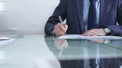 Businessman in a professional attire signing a contract with a pen on a modern glass desk in a...