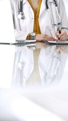Unknown doctor woman, wearing a stethoscope and yellow blouse under a lab coat, is using a clipboard and taking notes on glass desk in a medical office, close-up vertical view. Medicine concept