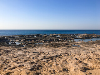 Along a sunny coastal stretch, rough stone and small tide pools dominate the foreground, while the wide blue sea and a clear sky form the backdrop, with a few people exploring the rocky edges.