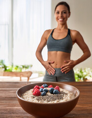 Femme sportive souriante d&rsquo;environ 30 ans touchant son ventre, devant un bol de porridge aux flocons d&rsquo;avoine, fruits rouges et graines de chia