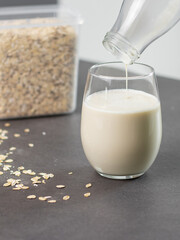 Close up of a female hand pouring oat milk into a glass on grey kitchen table. Vegan and non-dairy alternative milk