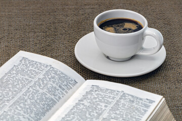 Black coffee in a white cup on a white saucer with a book and morning essentials on the table.
