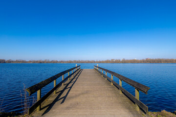 Late winter landscape, Wooden jetty (pier) extend into the Nieuwe meer (New lake) Beautiful sunny day with white fluffy cloud and blue sky background, Amsterdamse Bos (Forest) Amstelveen, Netherlands.