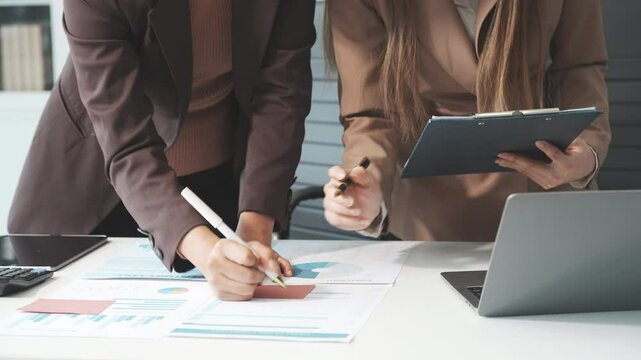 Two female business professionals in suits sit at desk, engaged in strategic discussion. They analyze financial planning, risk management, corporate growth while reviewing reports and market trends.