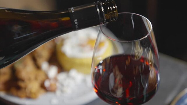A person is pouring wine into a glass. The wine is red and the glass is half full. The scene is set on a table with food, including a plate of food and a bowl. Scene is relaxed and social