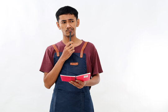 Asian barista man in blue apron taking order, writing on menu book list, thinking with pen on chin. Isolated image on white background - Powered by Adobe