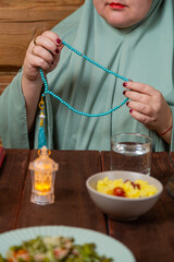 A woman in a light green hijab at a table with dates fingering her prayer beads reading