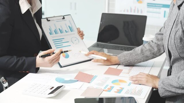 Two female business professionals in suits sit at desk, engaged in strategic discussion. They analyze financial planning, risk management, corporate growth while reviewing reports and market trends.