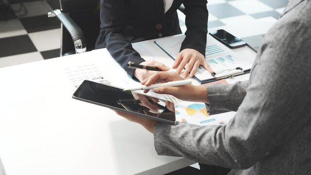Two female business professionals in suits sit at desk, engaged in strategic discussion. They analyze financial planning, risk management, corporate growth while reviewing reports and market trends.