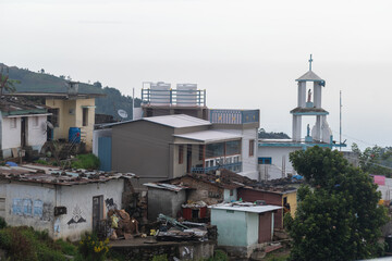 Obraz premium Nestled in Kodaikanal, the village showcases a blend of traditional and modern architecture. Houses are surrounded by lush greenery, while a church steeple rises against the morning sky.