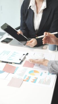 Two female business professionals in suits sit at desk, engaged in strategic discussion. They analyze financial planning, risk management, corporate growth while reviewing reports and market trends.